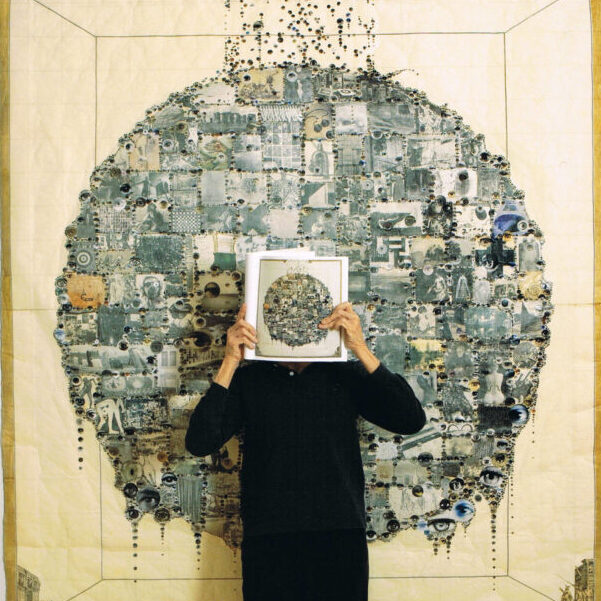 Portrait of Aldwyth standing in front of one of her large scale collages covering her face with a book.