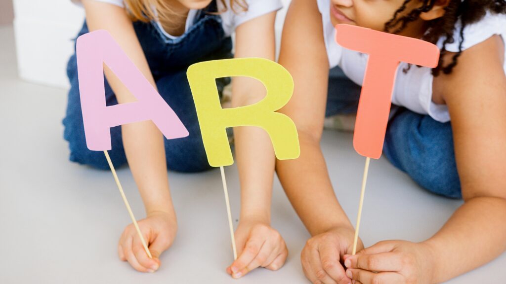 Photo of two children holding the letters A R T on sticks. Photo by Artem Podrez, through Pexels.